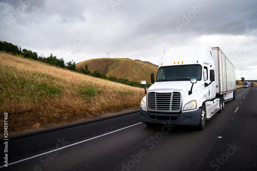 White big rig semi truck with dry van semi trailer driving in straight highway with hill roadside in California