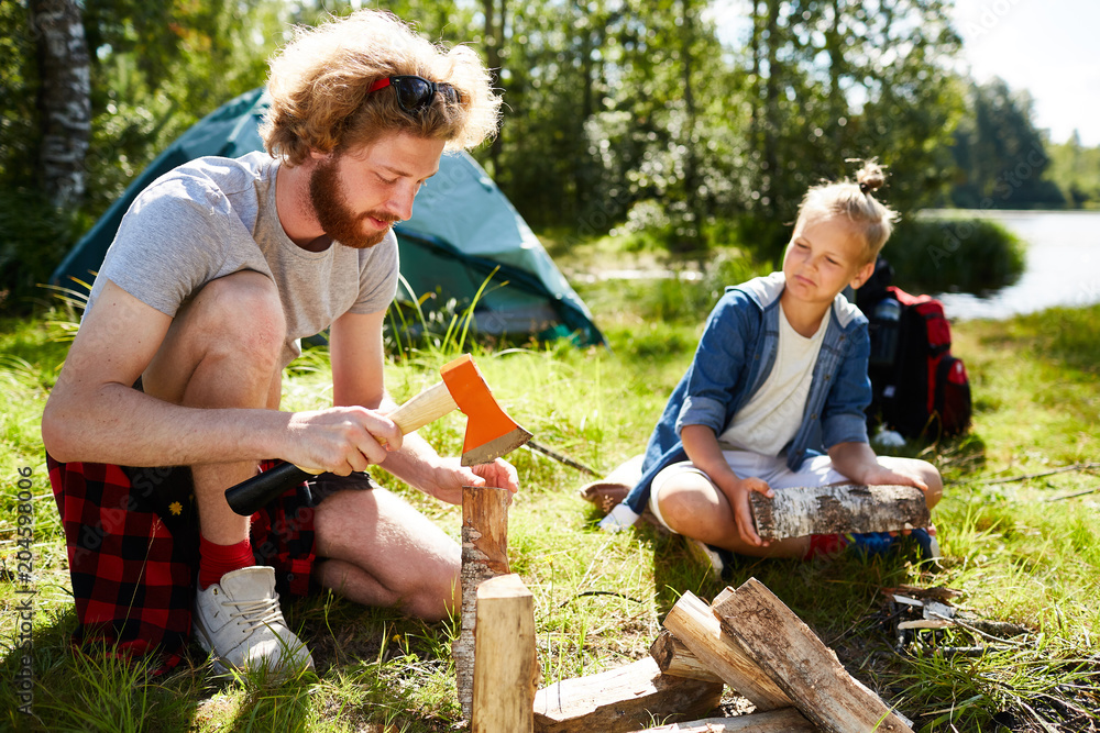 Young man with axe showing his son how to cut firewood for campfire on summer day during trip Young man with axe showing his son how to cut firewood for campfire on summer day during trip