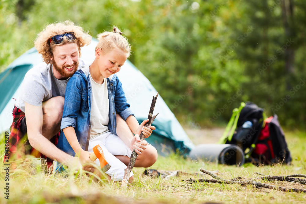 Young man and his son sharpening sticks with axe while going to prepare food on campfire Young man and his son sharpening sticks with axe while going to prepare food on campfire
