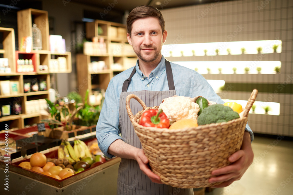 Fototapeta premium Happy supermarket assistant holding big basket with fresh vegetables during work