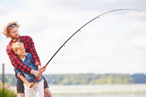 Little boy and his father holding fishing rod over lake while spending summer weekend in natural environment