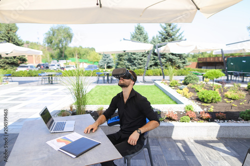 European businessman enjoying with virtual reality glasses, working with laptop and papers under beach umbrella near green plants. Concept of working on vacations and modern technology. photo