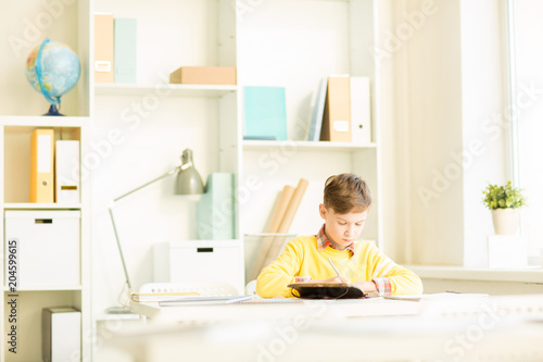 Cute young boy making notes or wiriting down home assignment in notebook while sitting by desk
