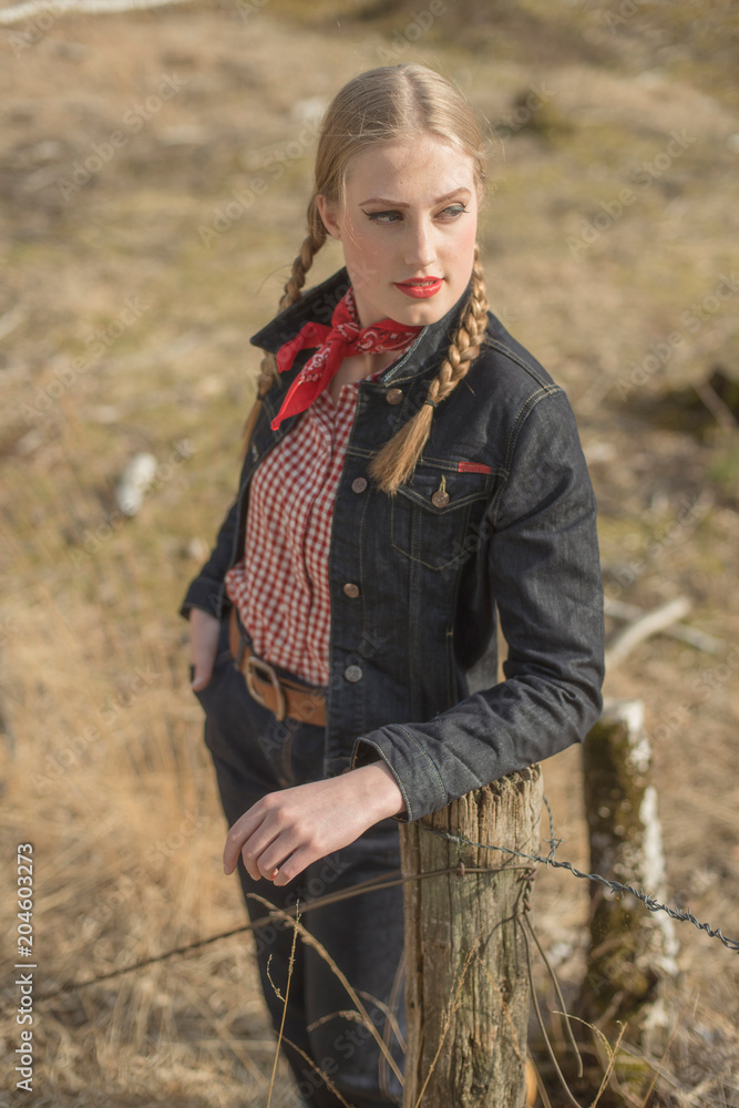 Retro 1950s blonde woman in jeans leaning against wooden post in field.