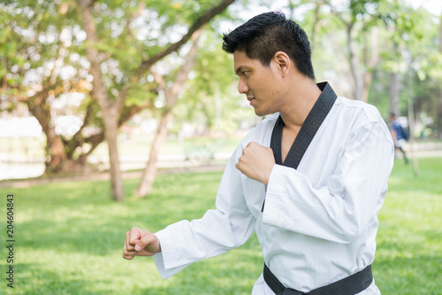 A male taekwondo is standing on green grass and looking ahead in public park, ready to fight.
