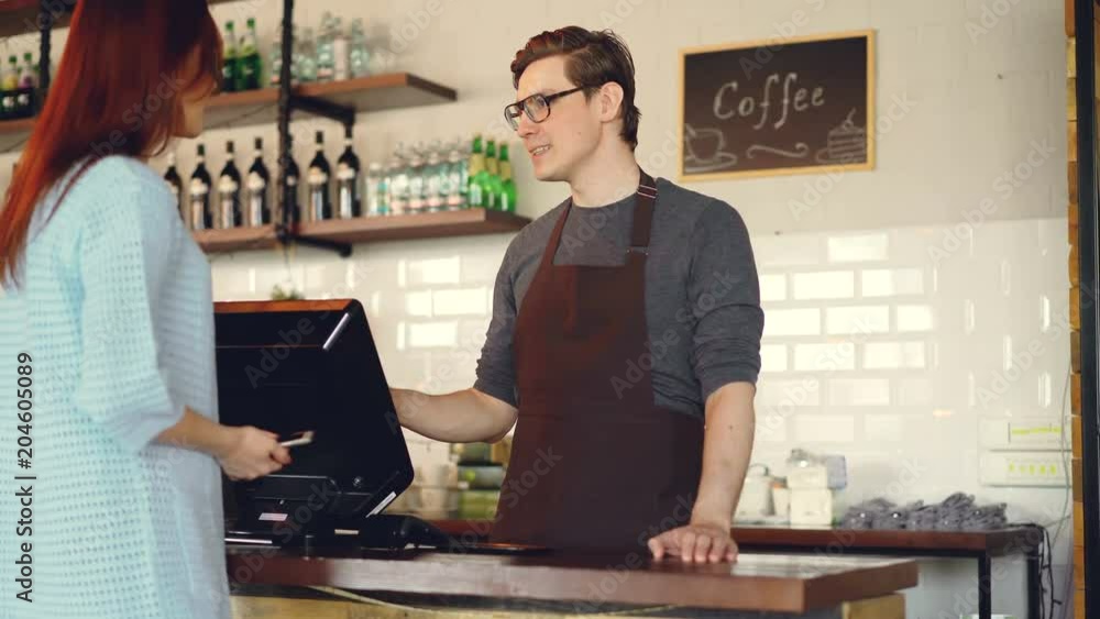 Pretty young lady is buying takeaway coffe in coffee-shop and paying with smartphone making contactless payment. Modern technology and banking concept. Pretty young lady is buying takeaway coffe in coffee-shop and paying with smartphone making contactless payment. Modern technology and banking concept.