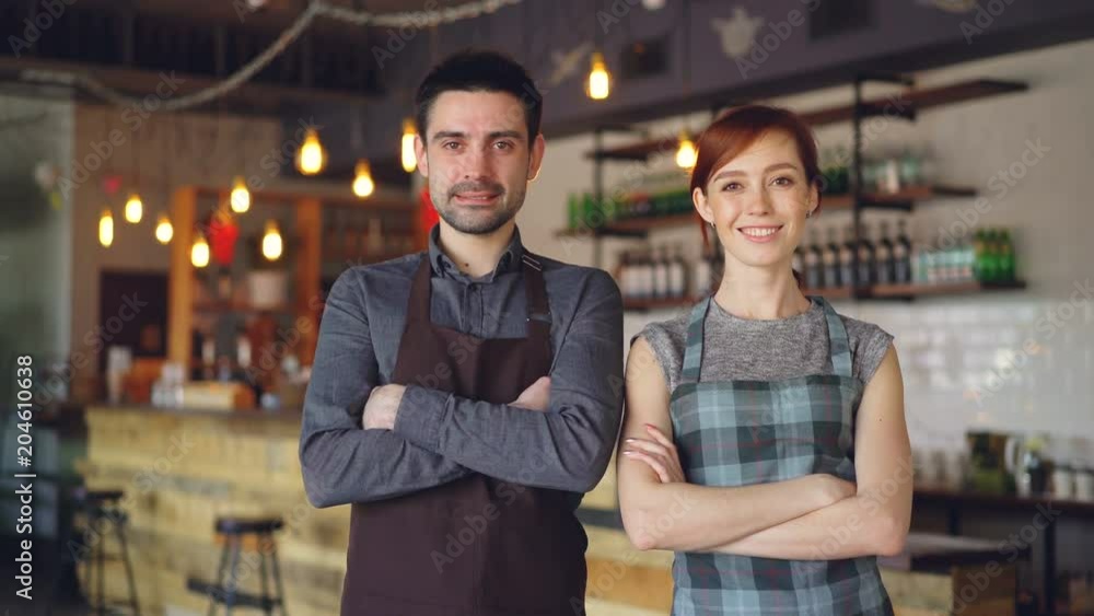 Portrait of two cheerful waiters standing inside cozy cafe, smiling and looking at camera. Successful business, happy attractive people and food service concept. Portrait of two cheerful waiters standing inside cozy cafe, smiling and looking at camera. Successful business, happy attractive people and food service concept.