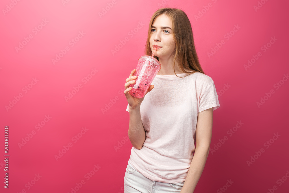 Portrait of beautiful young woman drinking fizzy drink from a bottle with a straw and looking at camera isolated on a pink background, is the text Portrait of beautiful young woman drinking fizzy drink from a bottle with a straw and looking at camera isolated on a pink background, is the text