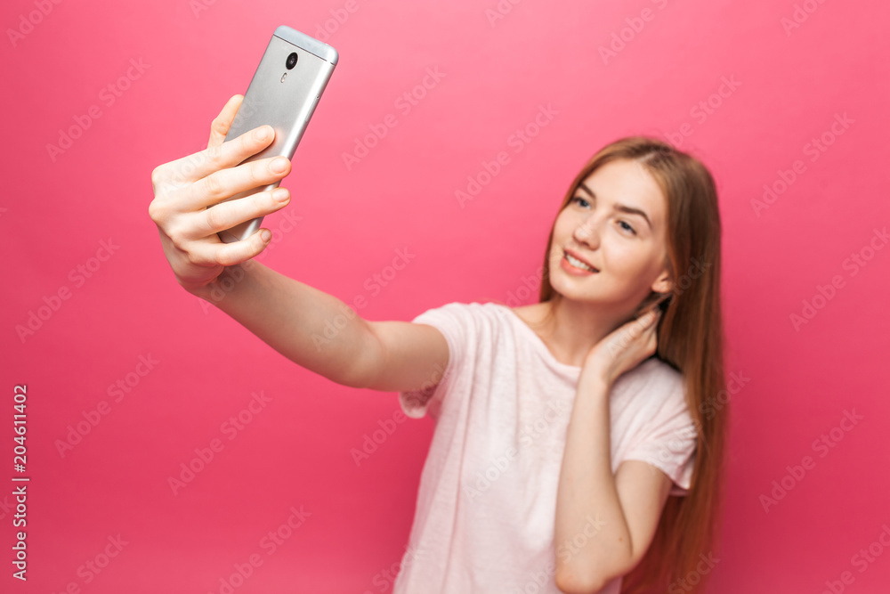 Portrait of cheerful beautiful girl, photographed, taking selfie, looking into the phone, funny enough, isolated on pink background, advertising Portrait of cheerful beautiful girl, photographed, taking selfie, looking into the phone, funny enough, isolated on pink background, advertising