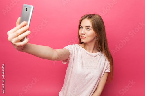 Portrait of cheerful beautiful girl, photographed, taking selfie, looking into the phone, funny enough, isolated on pink background, advertising