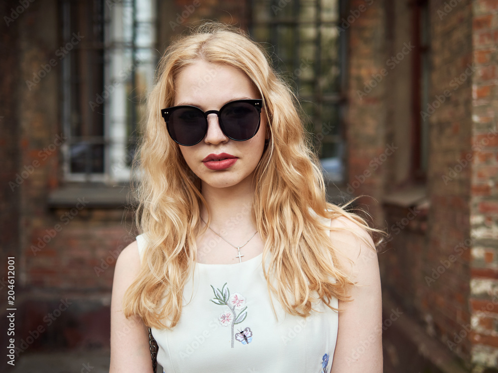 Close up of blonde student girl in trendy sunglasses posing against brick wall, enjoying warm sun light, recreation and relaxation on spring holiday Close up of blonde student girl in trendy sunglasses posing against brick wall, enjoying warm sun light, recreation and relaxation on spring holiday