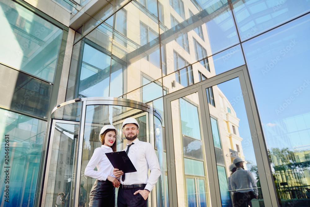 A girl with a man in construction white helmets and white shirts with drawings in hands against a glass office building. Engineers. Planning of construction A girl with a man in construction white helmets and white shirts with drawings in hands against a glass office building. Engineers. Planning of construction