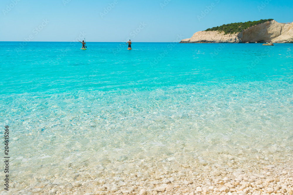A couple is floating on a their SUP boards into the turquoise waters of Porto katsiki in Lefkada Ionian island in Greece