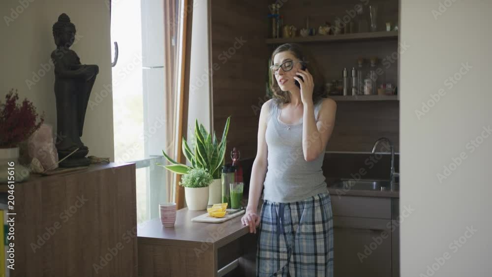 Attractive female in eyeglasses and pajamas standing in kitchen and talking on smartphone. Attractive female in eyeglasses and pajamas standing in kitchen and talking on smartphone.