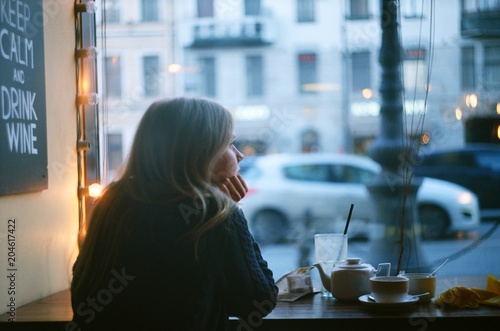 Lonely pensive blonde woman thinking in a coffee shop looking through the window at city street. evening