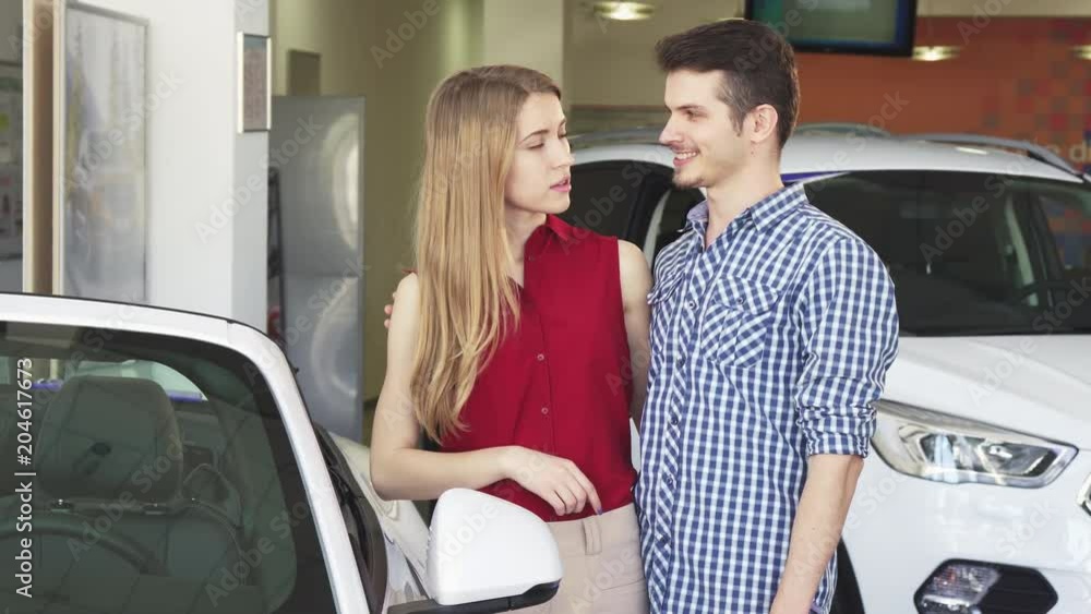 Attractive young happy couple hugging at the dealership salon while examining cars for sale. Handsome young man and his beautiful girlfriend choosing a new auto to buy. Attractive young happy couple hugging at the dealership salon while examining cars for sale. Handsome young man and his beautiful girlfriend choosing a new auto to buy.