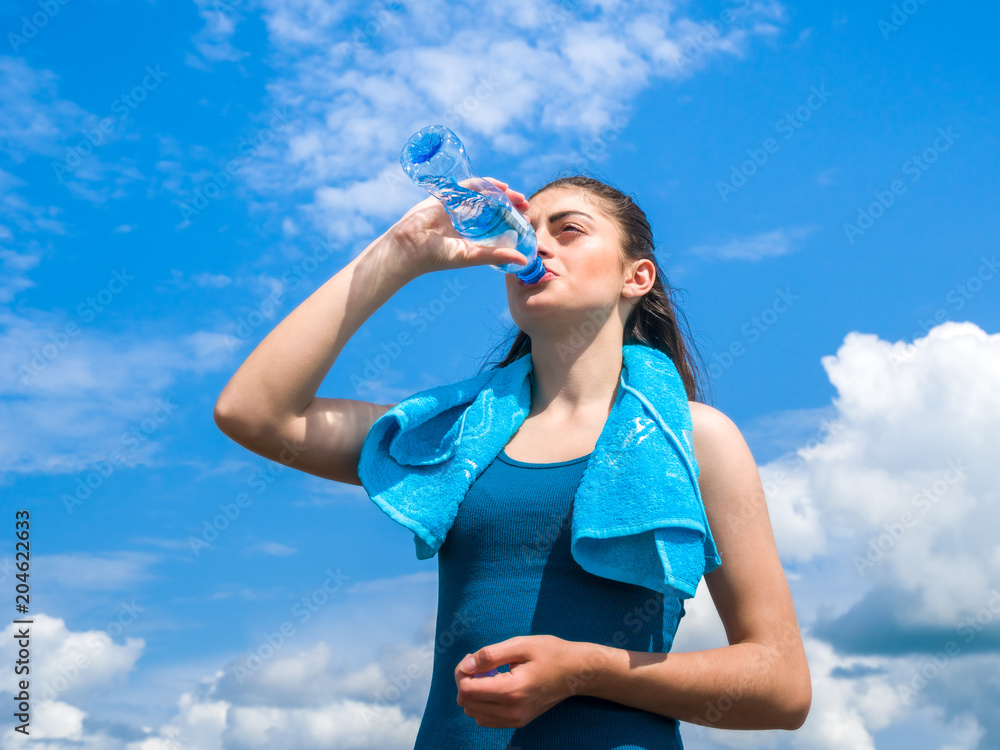 Beautifull girl runner is having break, drinking water against clear blue sky background Beautifull girl runner is having break, drinking water against clear blue sky background