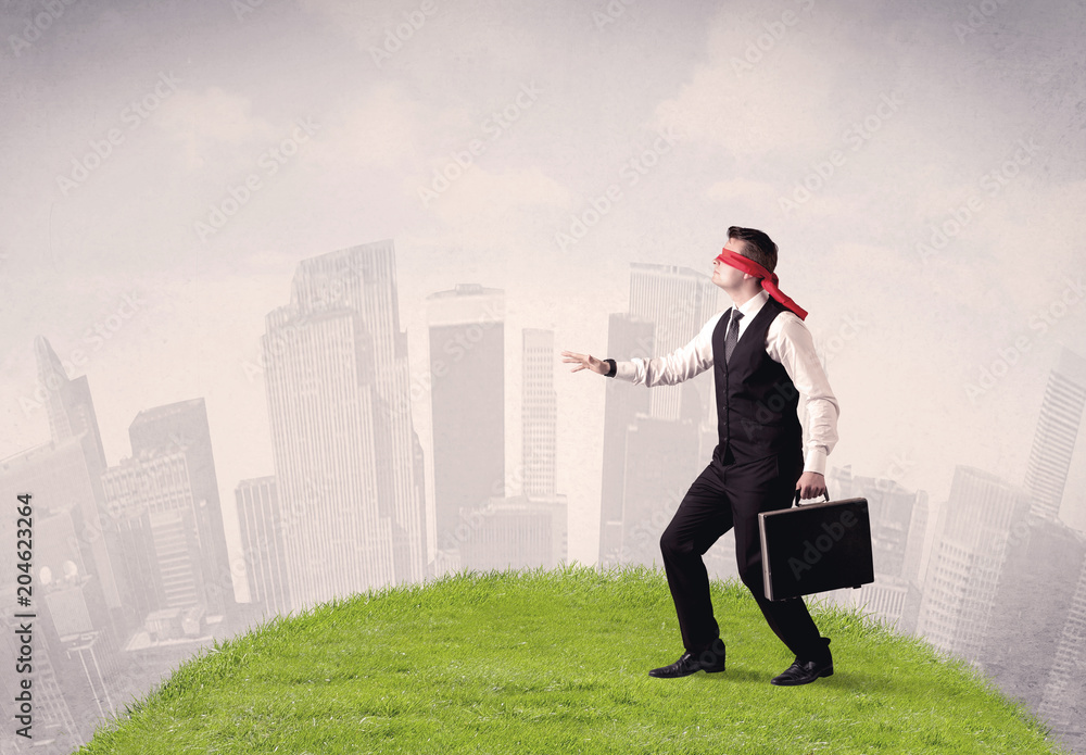 Young blindfolded businessman steps on a a patch of grass with a cloudy city in the background Young blindfolded businessman steps on a a patch of grass with a cloudy city in the background