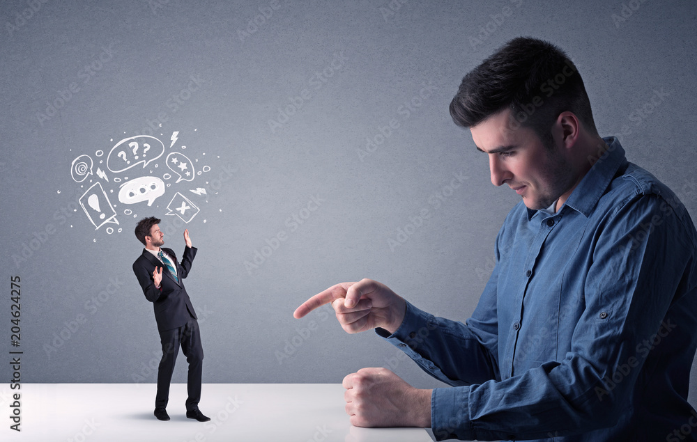 Young professional businessman being angry with an other miniature businessman who has drawn speech and thought bubbles above his head Young professional businessman being angry with an other miniature businessman who has drawn speech and thought bubbles above his head