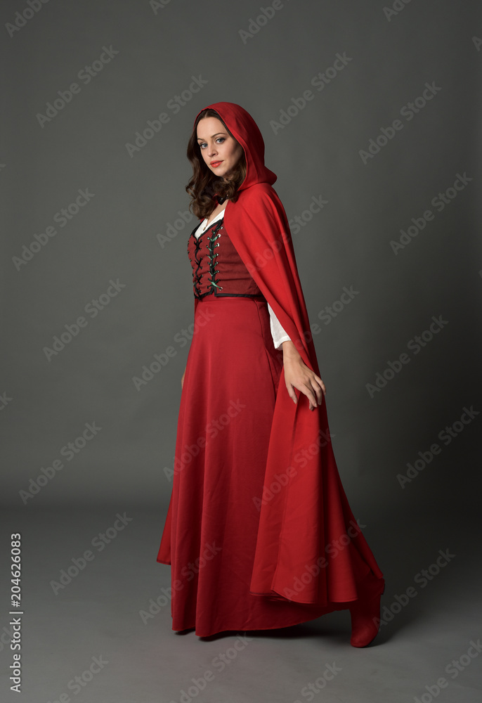 full length portrait of woman wearing red fantasy costume with cloak, standing pose on grey studio background.