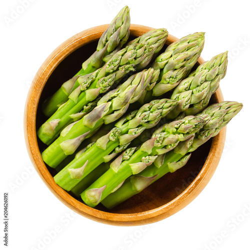 Fresh green asparagus tips in wooden bowl. Sparrow grass shoots. Cultivated Asparagus officinalis. Vegetable with thick stems and closed buds. Isolated macro food photo close up from above over white.