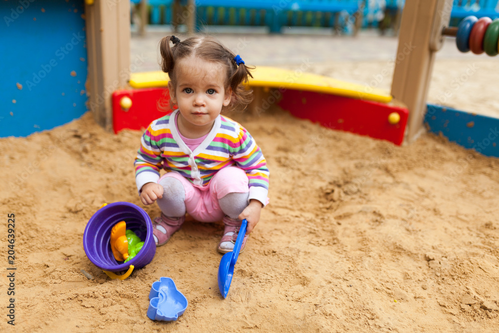 a little girl with two tails is dressed in a striped colorful jacket is playing in the sandbox on the playground a little girl with two tails is dressed in a striped colorful jacket is playing in the sandbox on the playground