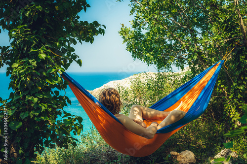 Rest. Man on vacation. The young man lies in a bright blue and orange hammock amidst the greenery of the sea shore and watches the beautiful scenery of the wild nature. The horizon is the sunny ocean.