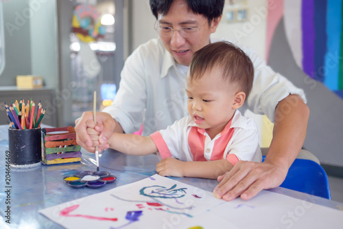 Cute smiling little Asian 18 months   1 year old toddler baby boy child painting with brush and watercolors  Businessman father painting with son after working time  Creative play for toddlers concept