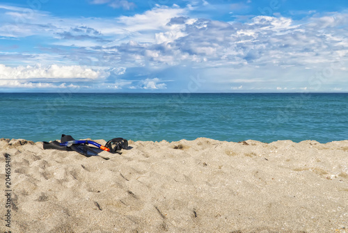 a mask for scuba diving, fins and a tube for breathing under the water on a sandy beach, against the background of the ocean and the blue sky in the clouds