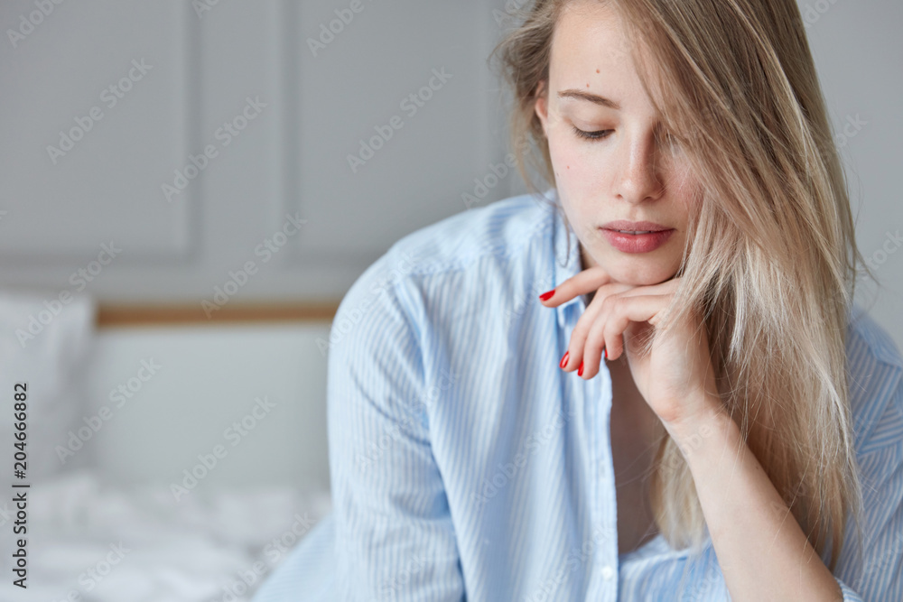 Close up shot of adorable woman holds chin, looks pensively down, poses against blurred bedroom interior, has healthy skin. Beauty, youth and rest concept. Relaxed female on comfortable bed. Close up shot of adorable woman holds chin, looks pensively down, poses against blurred bedroom interior, has healthy skin. Beauty, youth and rest concept. Relaxed female on comfortable bed.