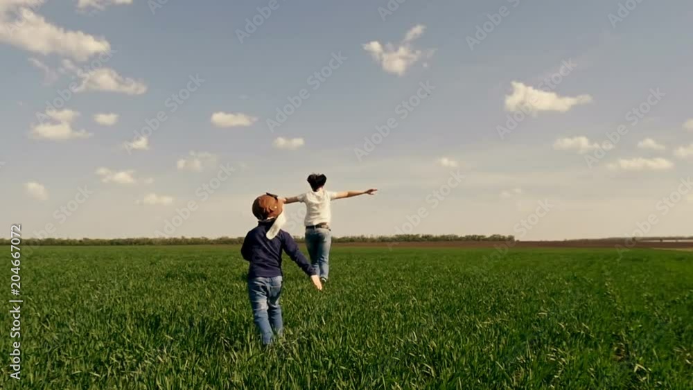 Happy mother and child running across the green field at sunset. Mom and her son are running along the green lawn in slow motion, showing the flight of the plane Happy mother and child running across the green field at sunset. Mom and her son are running along the green lawn in slow motion, showing the flight of the plane
