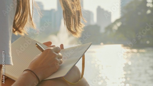 Young woman sitting on the bench in park and writing in diary, close-up photo