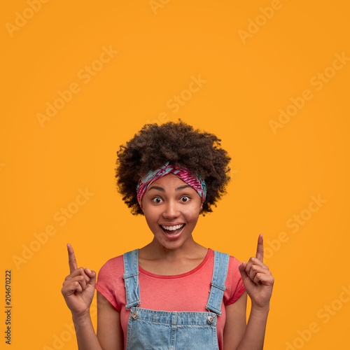 Happy African female with pleasant smile and excited look advertises something, indicates upwards on ceiling at blank copy space for your promotional text or advertisement. Hey, look there please