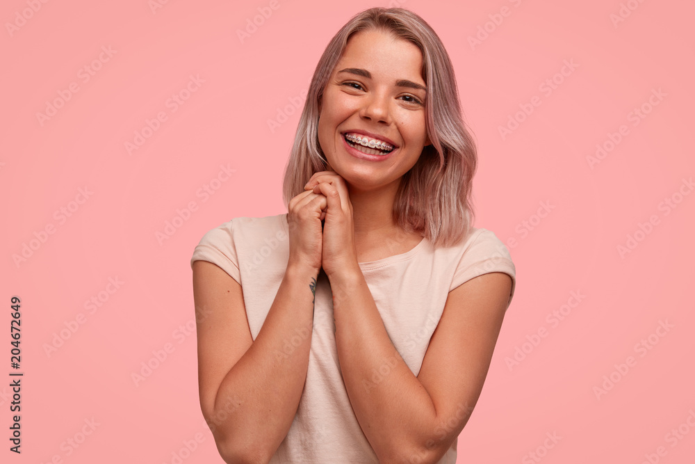 Fototapeta premium Horizontal shot of good looking female with positive smile, keeps hands together, being in good mood, rejoices winning contest, wears teeth braces, isolated over pink background. Happiness concept