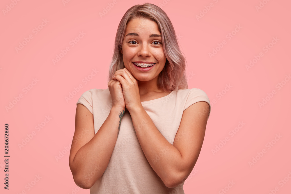 Pleasant looking happy cute female student rejoices successful graduation from university, smiles happily, wears braces and casual t shirt in one colour with background. Monochrome. Happiness Pleasant looking happy cute female student rejoices successful graduation from university, smiles happily, wears braces and casual t shirt in one colour with background. Monochrome. Happiness