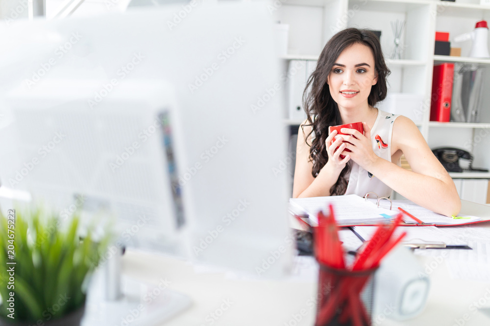 Beautiful young girl sits at office desk, looks at computer screen and holds a red mug in hands. Beautiful young girl sits at office desk, looks at computer screen and holds a red mug in hands.