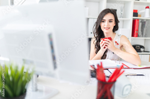 Beautiful young girl sits at office desk, looks at computer screen and holds a red mug in hands.