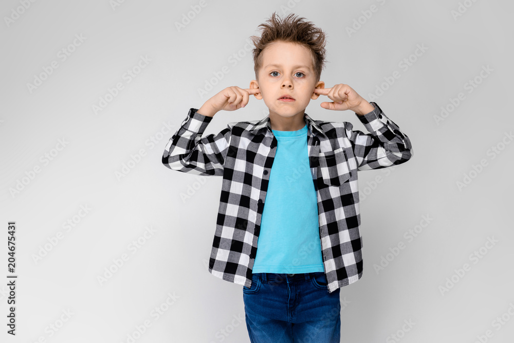 A handsome boy in a plaid shirt, blue shirt and jeans stands on a gray background. The boy folded his arms over his chest. The boy covered his ears with his fingers A handsome boy in a plaid shirt, blue shirt and jeans stands on a gray background. The boy folded his arms over his chest. The boy covered his ears with his fingers