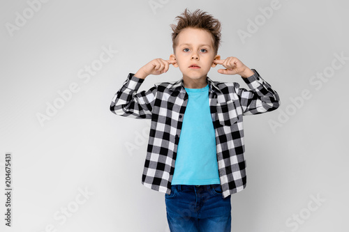 A handsome boy in a plaid shirt, blue shirt and jeans stands on a gray background. The boy folded his arms over his chest. The boy covered his ears with his fingers
