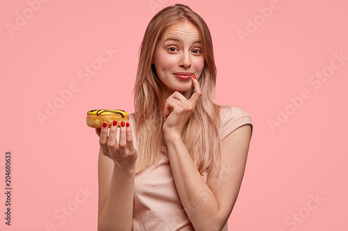 Charming young lovely female holds sweet doughnut, proposes you to eat delicious dessert, can`t refuse tasty eating, isolated over pink background. Gorgeous cute woman with donut. Junk food.