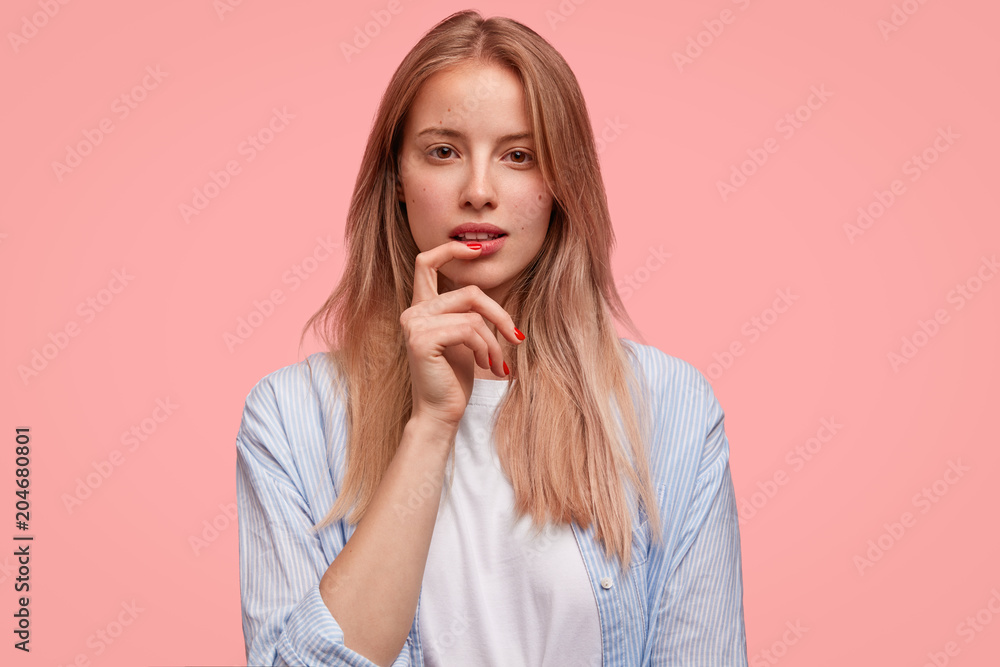 Lovely attractive female model with no make up, rests at home, thinks about how spend holidays, poses against pink studio background. European young woman looks thoughtfully and mysteriously Lovely attractive female model with no make up, rests at home, thinks about how spend holidays, poses against pink studio background. European young woman looks thoughtfully and mysteriously
