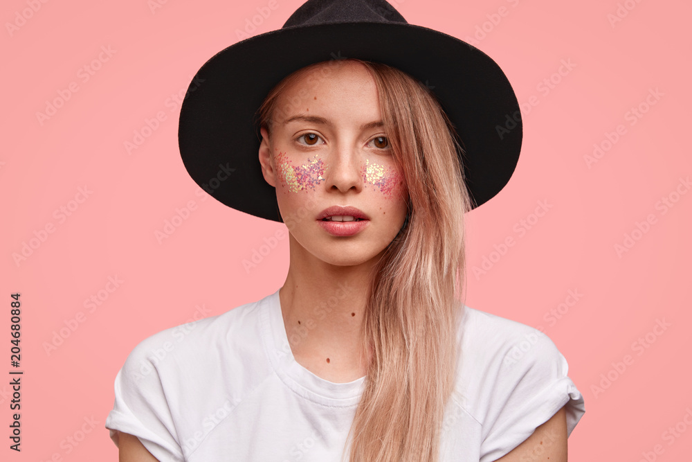 Portrait of delighted female model with glitter make up, wears elegant black hat and casual t shirt, poses against pink background. Serious beautiful woman with creative make up, stands indoor Portrait of delighted female model with glitter make up, wears elegant black hat and casual t shirt, poses against pink background. Serious beautiful woman with creative make up, stands indoor