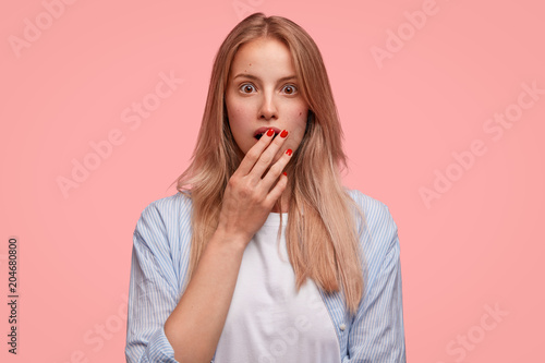 Studio shot of shocked Caucasian female covers mouth in terror, wears casual striped shirt, finds out about accident, poses against pink background. Lovely young woman recieves shocking news