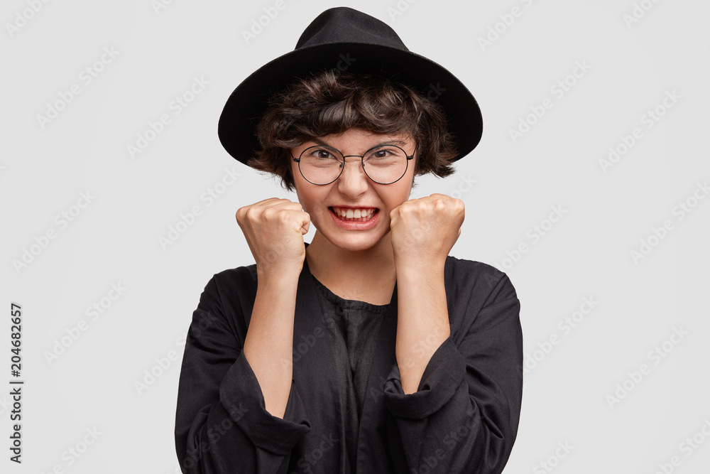 Irritated beautiful mixed race female shop assistant wears fashionable black hat, clenches fists, being annoyed with boring customers, expresses negativity and annoyance, uses gestures, isolated Irritated beautiful mixed race female shop assistant wears fashionable black hat, clenches fists, being annoyed with boring customers, expresses negativity and annoyance, uses gestures, isolated