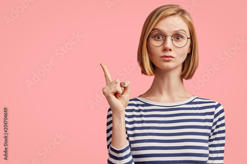 Portrait of serious stylish girl raises finger up, says: Attention please! Young stylish female teacher tries to explain something to pupils, has serious confident look, isolated on pink background