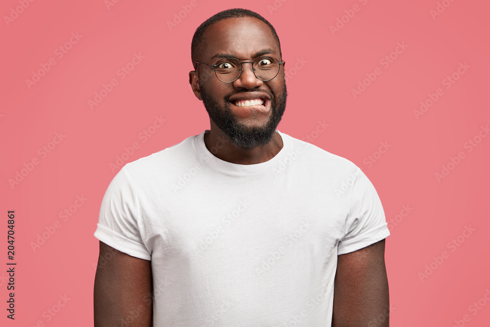 Isolated shot of annoyed black male bites lips in dissatisfaction, feels frustration and puzzlement, wears casual white t shirt, isolated over pink ackground. Neurotic African American man indoor Isolated shot of annoyed black male bites lips in dissatisfaction, feels frustration and puzzlement, wears casual white t shirt, isolated over pink ackground. Neurotic African American man indoor
