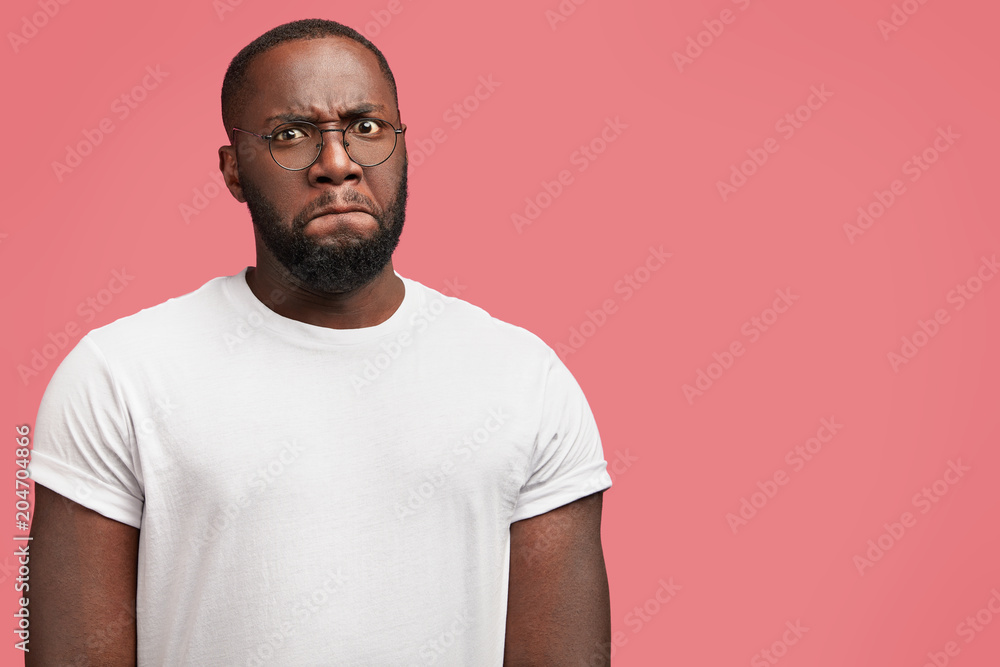 Horizontal shot of attractive black plump young male manager, tries to control his emotions, purses lips and looks angrily at camera, being annoyed with something, poses against pink background Horizontal shot of attractive black plump young male manager, tries to control his emotions, purses lips and looks angrily at camera, being annoyed with something, poses against pink background