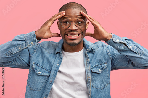 Canvas Print Sad depressed handsome young African American male clenches teeth, restrains tears, feels remorse, being in stressful situation, wears denim shirt, poses against pink background, feels pain