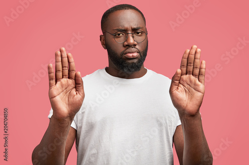 Middle aged black male shows stop gesture, keeps palms in front, has serious expression, wears casual t shirt, isolated on pink background. That`s forbidden! African American man rejects something photo