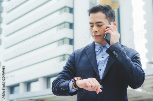 portrait attractive handsome business man using smartphone talking and looking smart watch at outdoor city with building background. technology connecting social online .